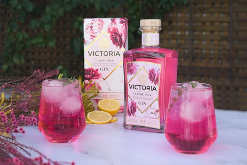 A bottle of Victoria Classic Pink Gin is placed on a marble surface, surrounded by floral decorations and slices of lemon. Two glasses filled with pink gin and ice cubes are garnished with herbs. The background features a wooden lattice and green foliage, giving a fresh and inviting atmosphere.