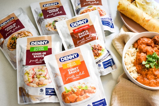 A selection of Denny sauce packets is displayed alongside a bowl of curry with rice and a rolled flatbread on a plate. The packets are flavors like Chicken A La King, Curry Sauce, and Stroganoff, among others. The curry in the bowl has a rich orange sauce garnished with fresh coriander.