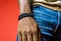 A close-up of a person's hand wearing two black bead bracelets. The background is a solid red color, and the person is wearing blue jeans and a light brown shirt.