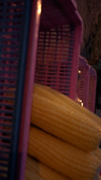 Several ears of yellow corn are stacked in front of a set of purple plastic crates. The scene suggests a market or a storage area with natural lighting and an emphasis on rustic textures.