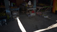A dimly lit market stall featuring wooden tables and crates filled with bananas and other produce. The concrete floor is partially illuminated by sunlight streaming in from the side. The stall contains various baskets and some bags of goods along the back wall.