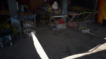 A dimly lit market stall featuring wooden tables and crates filled with bananas and other produce. The concrete floor is partially illuminated by sunlight streaming in from the side. The stall contains various baskets and some bags of goods along the back wall.
