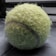 A worn and fuzzy tennis ball with a visible seam is placed on a dark surface. The texture of the ball appears aged and slightly frayed, indicating extensive use.