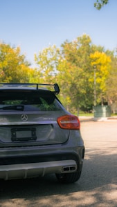 A luxury car with an AMG badge is parked in a sunny outdoor setting. The vehicle has a sleek design with visible chrome details and a rear spoiler. Lush, green trees with some yellow foliage are in the background under a clear blue sky.