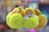 A collection of fluffy keychains, each featuring a doll face wearing a knitted yellow outfit. The focus is on the vibrant, furry pom-poms surrounding the doll faces. The background is blurred, enhancing the bright colors of the keychains.