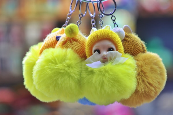 A collection of fluffy keychains, each featuring a doll face wearing a knitted yellow outfit. The focus is on the vibrant, furry pom-poms surrounding the doll faces. The background is blurred, enhancing the bright colors of the keychains.