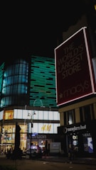 A large illuminated storefront at night with various retail signs including Macy's, H&M, Verizon, and Sunglass Hut. The area is well-lit with modern architectural elements and features neon lights, large glass windows, and busy street elements.