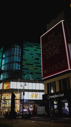 A large illuminated storefront at night with various retail signs including Macy's, H&M, Verizon, and Sunglass Hut. The area is well-lit with modern architectural elements and features neon lights, large glass windows, and busy street elements.
