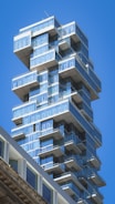 A modern high-rise building featuring an unusual stacked design with multiple glass balconies protruding at various levels against a clear blue sky.