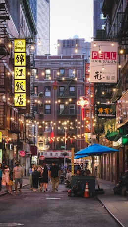 A vibrant urban street scene with colorful neon signs in an Asian language hanging on buildings. Festive string lights are draped overhead, enhancing the lively atmosphere. A mix of people are walking on the street and gathered at an outdoor seating area under a blue canopy. Buildings show varied architecture against a backdrop of taller city structures.