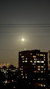 A nighttime cityscape with a full moon shining brightly in a dark sky above a residential building. Electric power lines stretch across the sky. The building features illuminated windows, suggesting occupancy, and additional buildings with lights can be seen in the background.