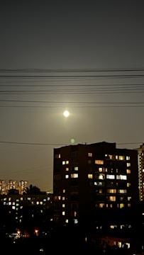 A nighttime cityscape with a full moon shining brightly in a dark sky above a residential building. Electric power lines stretch across the sky. The building features illuminated windows, suggesting occupancy, and additional buildings with lights can be seen in the background.