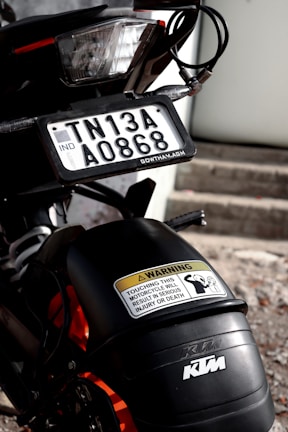 A close-up view of a motorcycle with a focus on the rear section. The motorcycle features a license plate with the registration number TN13A A0868, an Indian registration identifier. Above the rear wheel is a warning sticker indicating that touching the motorcycle can lead to serious injury or death. The vehicle branding 'KTM' is visible on the bodywork.