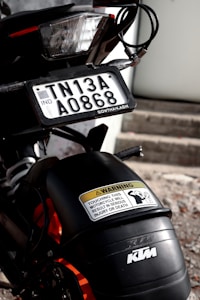 A close-up view of a motorcycle with a focus on the rear section. The motorcycle features a license plate with the registration number TN13A A0868, an Indian registration identifier. Above the rear wheel is a warning sticker indicating that touching the motorcycle can lead to serious injury or death. The vehicle branding 'KTM' is visible on the bodywork.