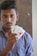 A young man with short, dark hair and a checkered shirt holds three playing cards in his hand. The cards are the Ace of Spades, a Joker, and the Ace of Hearts. He has a serious expression on his face.