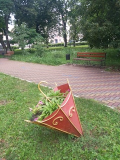 An outdoor park setting features a unique decorative planter shaped like an upside-down red umbrella with golden accents. Positioned on green grass, the planter holds an assortment of greenery and pink flowers. In the background, a paved pathway runs through the park, flanked by two wooden benches with metal frames. Tall trees with lush green foliage create a serene atmosphere.