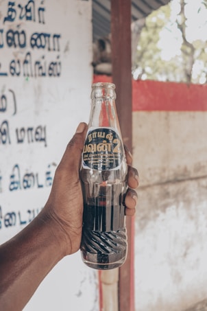 A hand holding a glass soda bottle with a dark liquid inside. The label on the bottle has writing in a script likely to be Tamil. The background includes a wall with text in the same script and some outdoor elements.