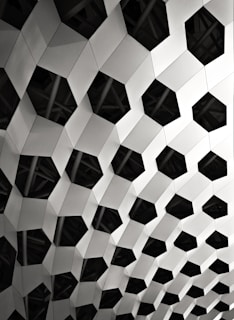 A geometric pattern composed of interconnected hexagonal shapes with a three-dimensional appearance. The structure appears metallic and features alternating light and dark surfaces, creating a play of light and shadow.