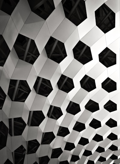 A geometric pattern composed of interconnected hexagonal shapes with a three-dimensional appearance. The structure appears metallic and features alternating light and dark surfaces, creating a play of light and shadow.