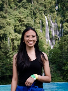 A young woman with long black hair and glasses standing in front of a lush green forest with a waterfall in the background. She is wearing a black tank top and blue pants and is smiling at the camera. There is a single light bulb hanging above her.