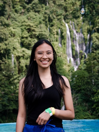 A young woman with long black hair and glasses standing in front of a lush green forest with a waterfall in the background. She is wearing a black tank top and blue pants and is smiling at the camera. There is a single light bulb hanging above her.