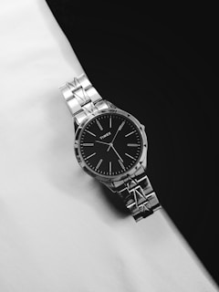 A silver wristwatch with a black face and minimalist design is positioned diagonally against a monochrome background. The watch has a metal band with visible links and a simple dial with slender hour markers and hands.