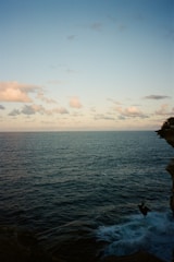 A vast ocean stretches towards the horizon under a partly cloudy sky. The horizon line is slightly elevated in the composition. On the right side, a figure is performing a bungee jump off a cliff into the blue waters below, creating a sense of adventure and exhilaration.