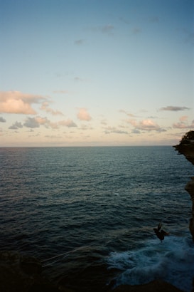A vast ocean stretches towards the horizon under a partly cloudy sky. The horizon line is slightly elevated in the composition. On the right side, a figure is performing a bungee jump off a cliff into the blue waters below, creating a sense of adventure and exhilaration.