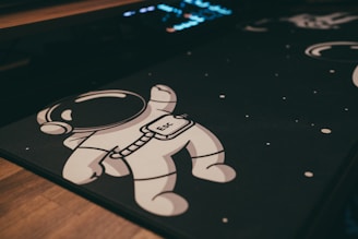 A cartoon-style astronaut is depicted on a dark surface with small white dots reminiscent of stars in space. The astronaut is in a playful pose, wearing a helmet, and has the word 'Esc' on its suit. The background includes a partially visible illuminated keyboard, adding a tech or gaming theme to the image.