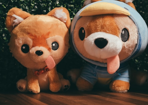 Two plush toys resembling dogs are placed side by side on a wooden floor with a backdrop of green foliage. The toy on the left has a simple design with a happy expression and a protruding tongue, while the one on the right wears a blue costume with a duck-bill hood.