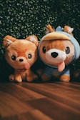 Two plush toys resembling dogs are placed side by side on a wooden floor with a backdrop of green foliage. The toy on the left has a simple design with a happy expression and a protruding tongue, while the one on the right wears a blue costume with a duck-bill hood.