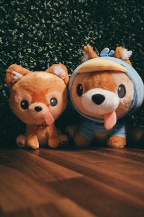 Two plush toys resembling dogs are placed side by side on a wooden floor with a backdrop of green foliage. The toy on the left has a simple design with a happy expression and a protruding tongue, while the one on the right wears a blue costume with a duck-bill hood.