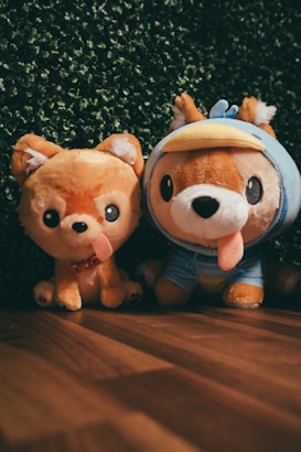 Two plush toys resembling dogs are placed side by side on a wooden floor with a backdrop of green foliage. The toy on the left has a simple design with a happy expression and a protruding tongue, while the one on the right wears a blue costume with a duck-bill hood.