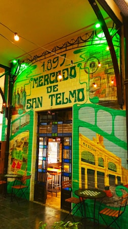 A colorful mural depicting the Mercado de San Telmo, featuring vivid shades of green and yellow. The architectural detail of the market entrance is highlighted, surrounded by artistic illustrations. String lights add a warm ambiance, while small tables and chairs are positioned in the foreground.