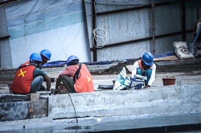 Several construction workers wearing blue helmets and orange safety vests are engaged in work on a concrete structure. They are surrounded by construction materials and tools. A partially constructed building or structure forms the background.
