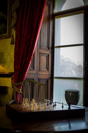 A cozy room features a chessboard set on a wooden table, positioned near a large window with sheer curtains allowing soft, diffused light. Rich, red velvet curtains frame the window, adding a touch of elegance alongside a decorative vase placed on the ledge. A wooden chair with an intricate backrest is tucked under the table.