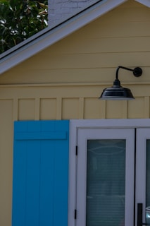 A section of a building featuring a yellow exterior wall with vertical paneling is depicted. It includes a blue wooden shutter next to a glass door framed in white. Above the door, a black outdoor light fixture is attached to the wall. In the background, part of a white brick chimney and green foliage can be seen.