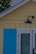 A section of a building featuring a yellow exterior wall with vertical paneling is depicted. It includes a blue wooden shutter next to a glass door framed in white. Above the door, a black outdoor light fixture is attached to the wall. In the background, part of a white brick chimney and green foliage can be seen.