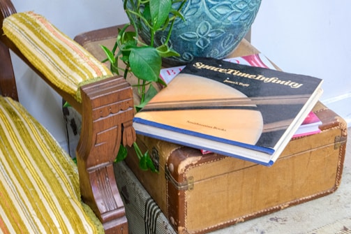 A vintage suitcase is used as a makeshift table, with a book titled 'SpaceTime Infinity' resting on it. The book cover features an image of the planet Saturn. Nearby is a potted plant with trailing green leaves, and a wooden chair with yellow and white striped upholstery is partially visible.