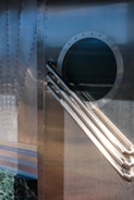 The image features a metallic structure with riveted panels and a circular, window-like element. Multiple cylindrical rods extend diagonally across the surface. The lower part of the image includes a section with a textured, dark green material and horizontal stripes.