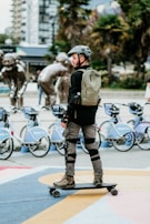 A person is skateboarding equipped with safety gear, including a helmet, pads, and a backpack. In the background, there's a lineup of rental bicycles and a series of metal sculptures depicting figures in motion. The scene is set in an urban environment with trees and buildings.
