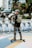 A person is skateboarding equipped with safety gear, including a helmet, pads, and a backpack. In the background, there's a lineup of rental bicycles and a series of metal sculptures depicting figures in motion. The scene is set in an urban environment with trees and buildings.