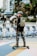 A person is skateboarding equipped with safety gear, including a helmet, pads, and a backpack. In the background, there's a lineup of rental bicycles and a series of metal sculptures depicting figures in motion. The scene is set in an urban environment with trees and buildings.