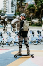 A person is skateboarding equipped with safety gear, including a helmet, pads, and a backpack. In the background, there's a lineup of rental bicycles and a series of metal sculptures depicting figures in motion. The scene is set in an urban environment with trees and buildings.