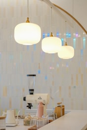 A modern kitchen counter features minimalistic decor with glass jars and a coffee grinder. Three pendant lights hang above, casting a warm glow. The background showcases glossy, iridescent tiles in neutral tones, adding texture and subtle color shifts.