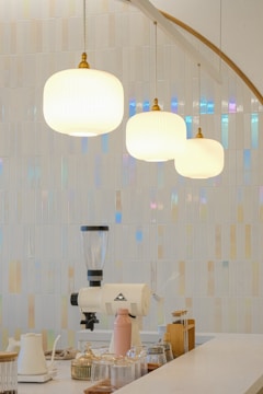 A modern kitchen counter features minimalistic decor with glass jars and a coffee grinder. Three pendant lights hang above, casting a warm glow. The background showcases glossy, iridescent tiles in neutral tones, adding texture and subtle color shifts.