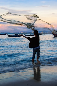 A person standing in shallow water is casting a large fishing net into the sea. The scene takes place during sunset, with soft pink and purple hues in the sky, and several boats floating in the background. Ripples form in the blue water as the net is thrown.