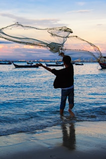 A person standing in shallow water is casting a large fishing net into the sea. The scene takes place during sunset, with soft pink and purple hues in the sky, and several boats floating in the background. Ripples form in the blue water as the net is thrown.