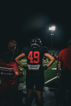 A football player stands on a field at night, wearing a black uniform with the number 49 in red on the back and the name Douglas visible. Stadium lights illuminate the scene, casting a dramatic glow. Two other people are nearby, one with long hair in a maroon shirt and another in a red shirt.