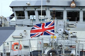 A naval ship with the Union Jack flag prominently displayed, surrounded by various equipment and lifebuoys. The vessel's structure includes windows and metallic surfaces.
